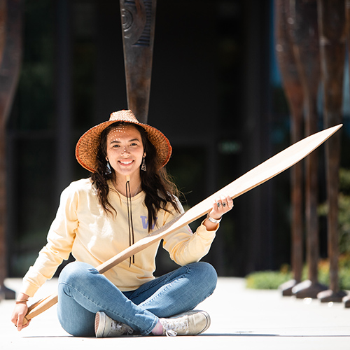 Stephanie Masterman in front of the Burke Museum, holding a cedar oar that she carved.