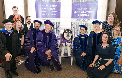 Department of Scandinavian Studies faculty, most in caps and gowns, flanking the UW Husky dog, Dubs, with a department banner behind them.