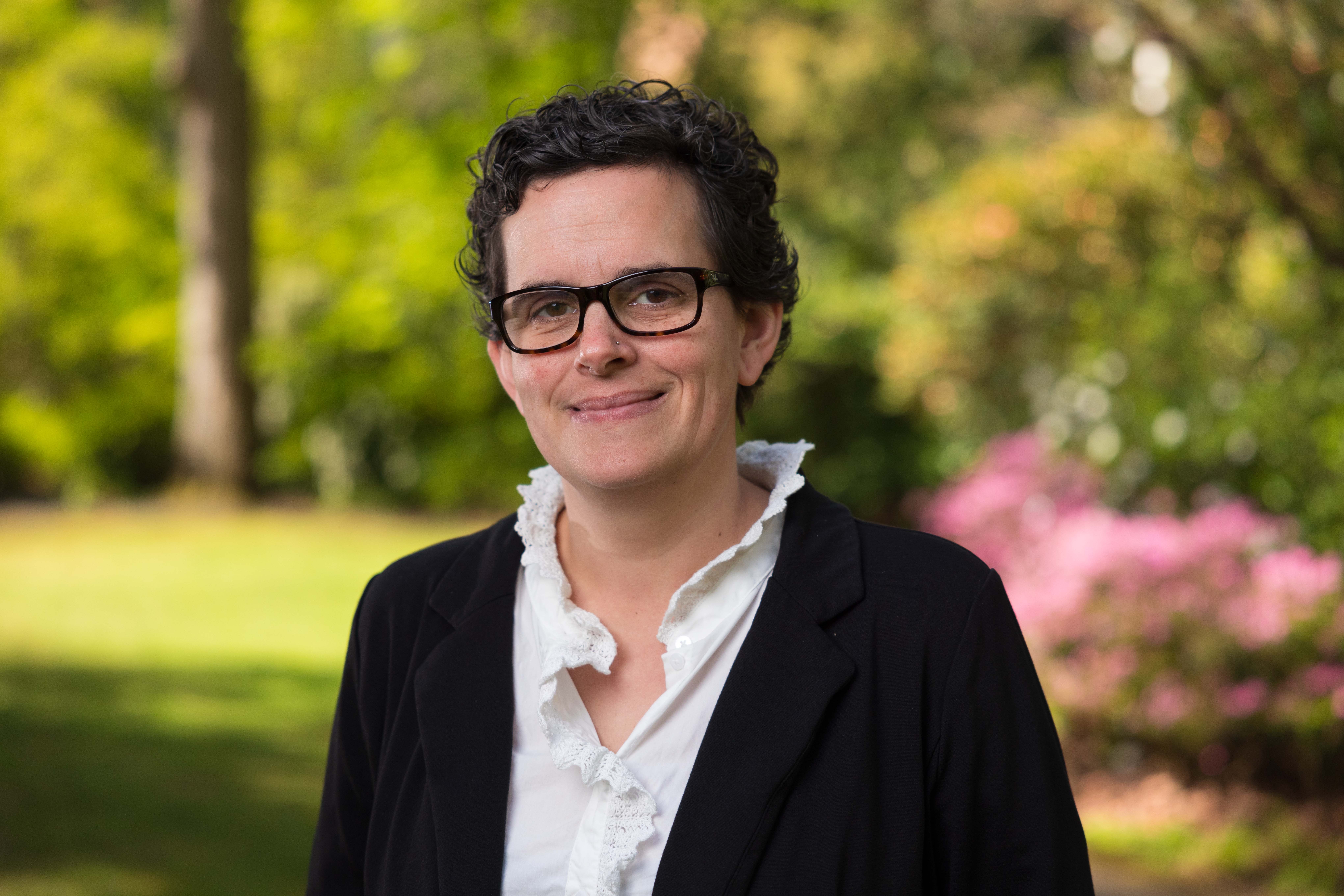 Headshot of Helen Oland wearing glasses and black and white top, set against a blurry tree lined background