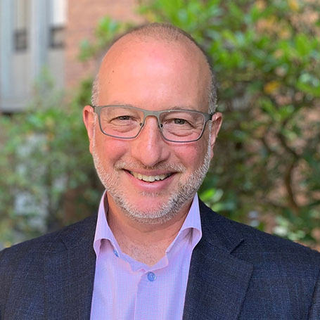 A smiling man with glasses posed in front of greenery.
