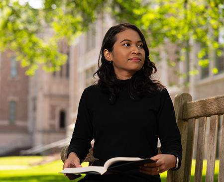 Anselma Prihandita on the UW campus, sitting on a bench with an open book in her lap.