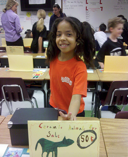 Young Kyra Wolfenbarger, in elementary school, in front of a handwritten sign that reads "Ceramic Animals for Sale, 50 cents"