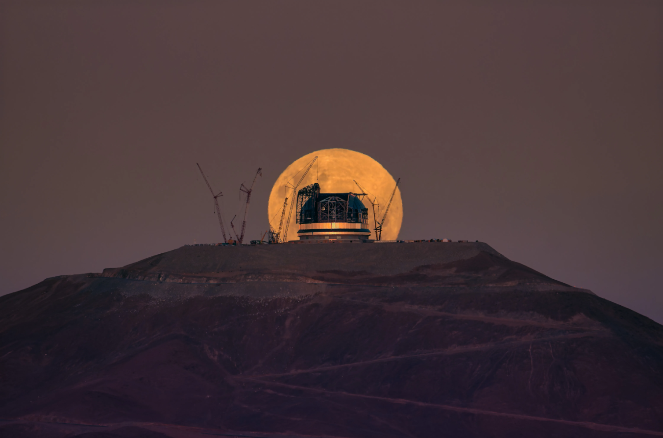 An orange Moon rising behind the partially built Extremely Large Telescope on the top of a hill.