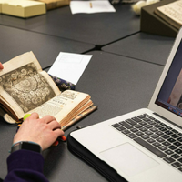 A person looking at a book and a laptop on a table