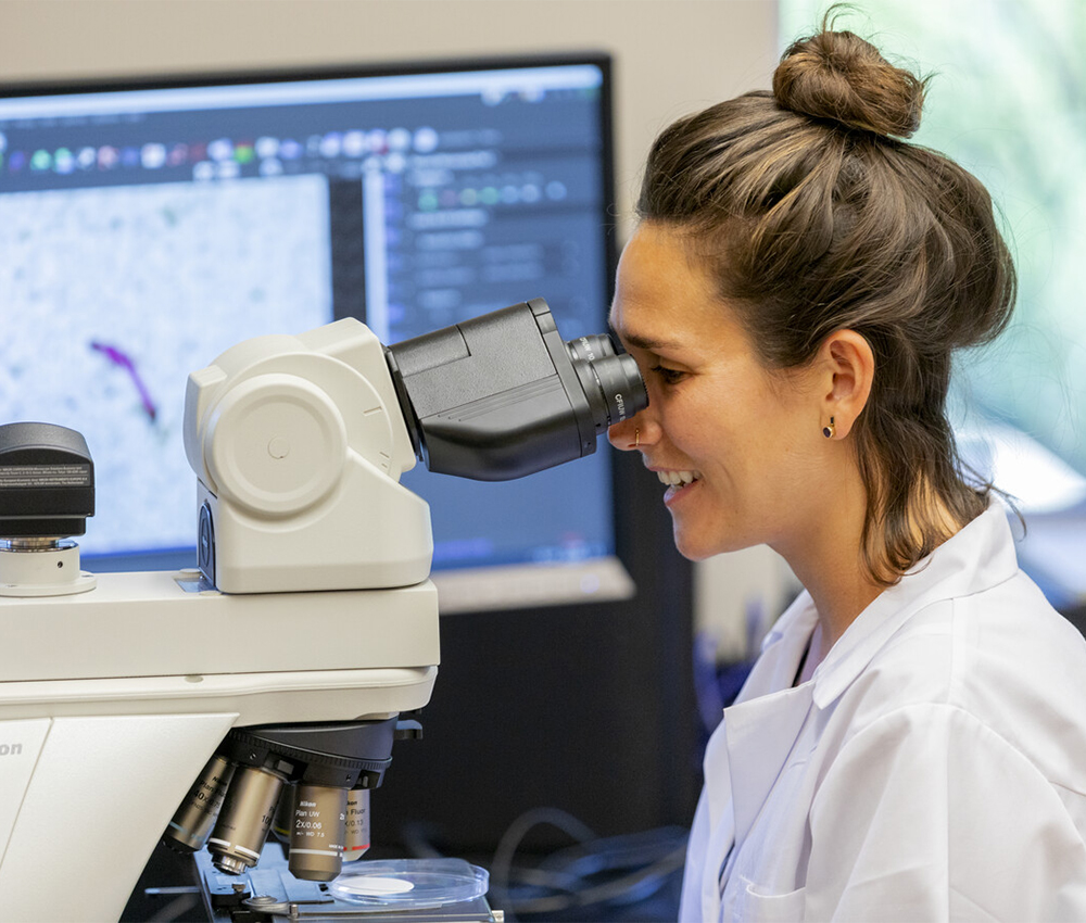 Person in lab coat smiles while looking into a microscope.