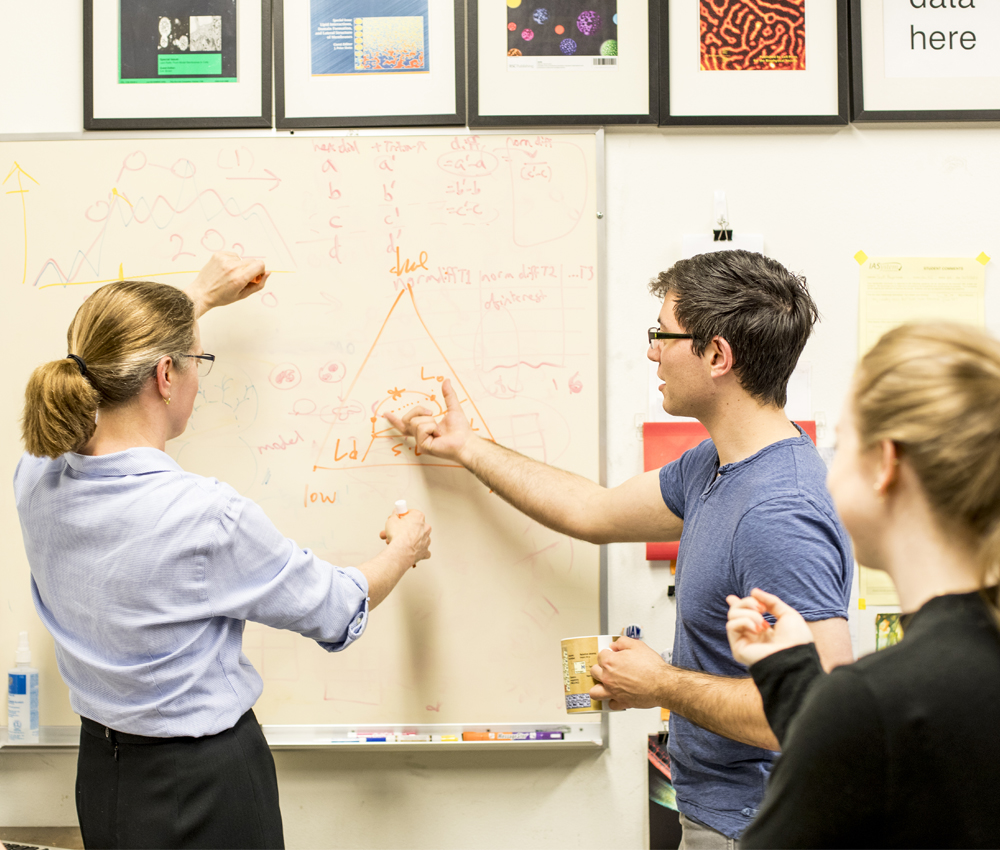 Three people work at a whiteboard with a variety of symbols and figures.