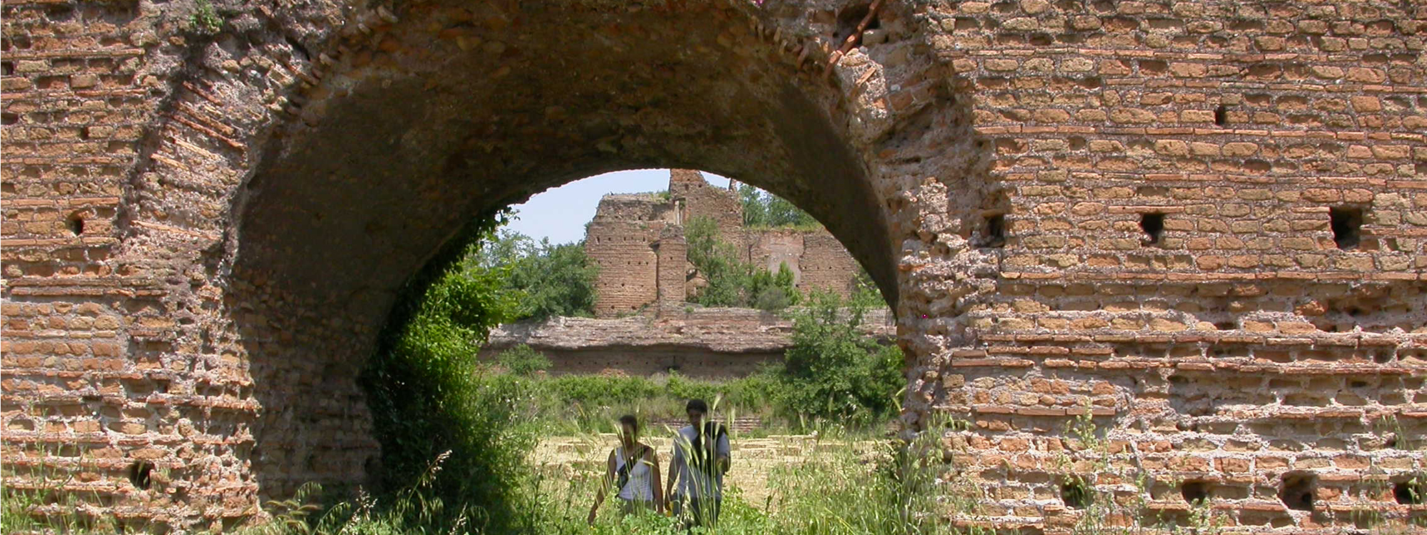 Two people walk under a brick arch in some ruins.