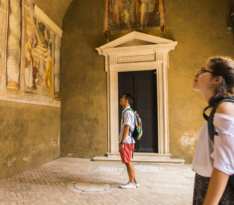 Two people wearing backpacks look up at art in an ancient Greek style building.