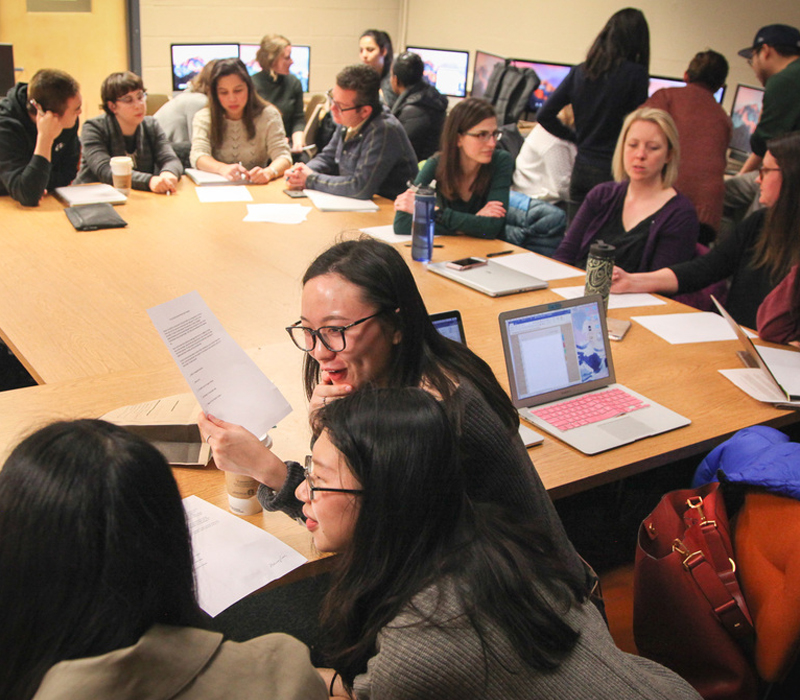 A large group of students talk with each other at a table with laptops and papers.