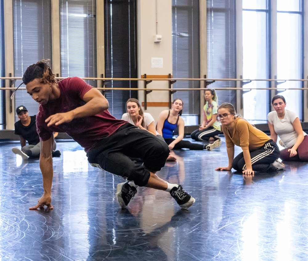 Students sit and watch someone demonstrate a dance move in a studio.