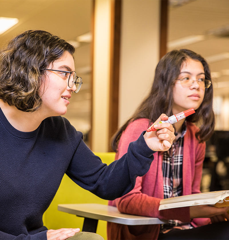 Two students are participating in a class discussion, with one pointing at the front of the room.