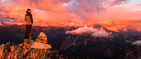 A student stands on the peak of a mountain looking out at sunset with pink and orange clouds.