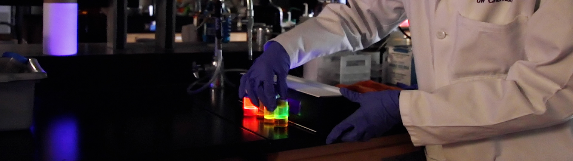 Chemistry student in dark lab with colorful vials lit up by UV light