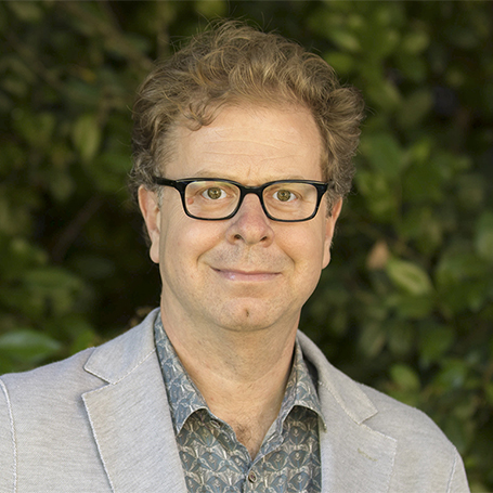 George Lovell in button down shirt and jacket standing in front of dark green bushes