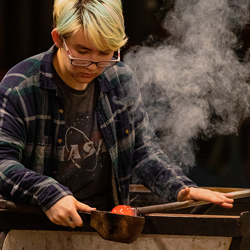 Glassblowing student working on piece, with smoke rising from it