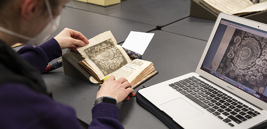 Student looking at an old book, with image from the book on the laptop next to her. 