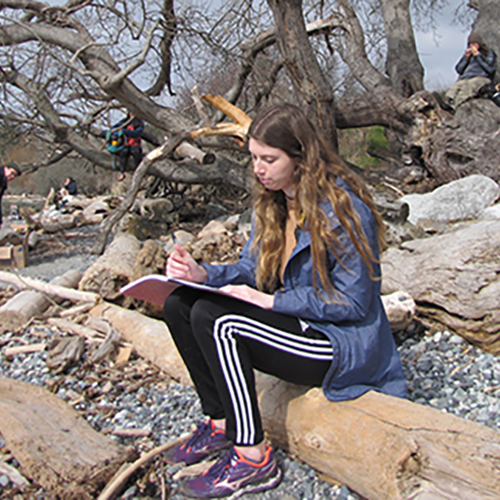 Student writing in notebook, sitting along a rocky shoreline