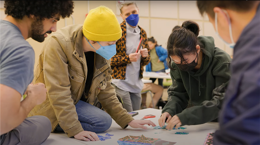 Students around a table with colorful shapes, collaborating on an art project