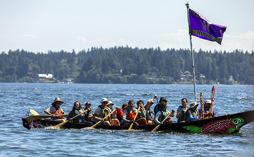 UW canoe on the water during the Tribal Canoe Journey, with UW flag above it, waving in the breeze