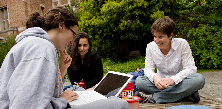 Three students seated outside talking while one looks at laptop.