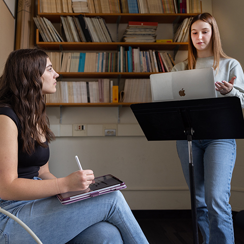 A seated student consultant takes notes while another student stands to practice a speech.