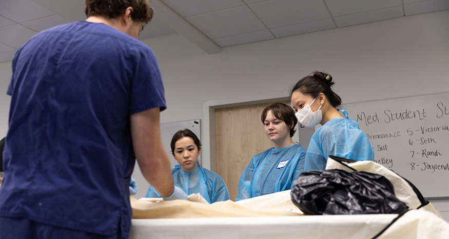 Three students look at an unseen object on a table in the anatomy lab. 