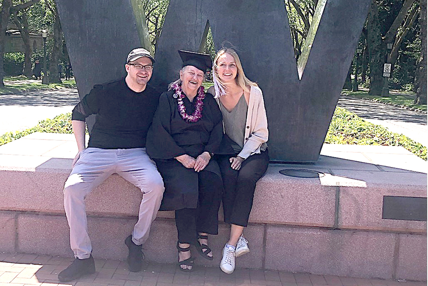 Linda Allen seated in front of the large "W" sign at the entrance to campus, flanked by a grandson and granddaughter, both young adults