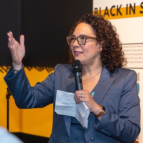 Ralina Joseph holding a microphone and speaking, with an Interrupting Privilege display panel behind her, at the Northwest African American Museum.