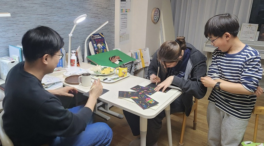 Katie Ruesink, Hojun, and his father sit around a table working on an art project.