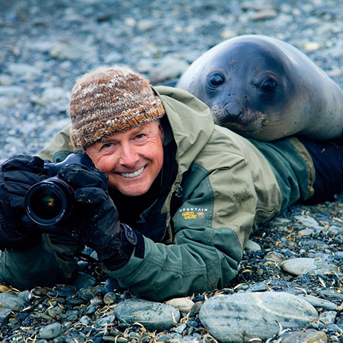 Art Wolfe laying on the ground holding his camera, with a seal perched on top of his legs