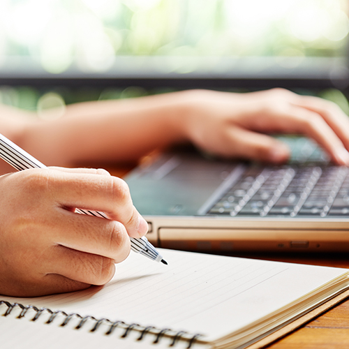 Person with one hand on a laptop keyboard, the other writing in a notebook.