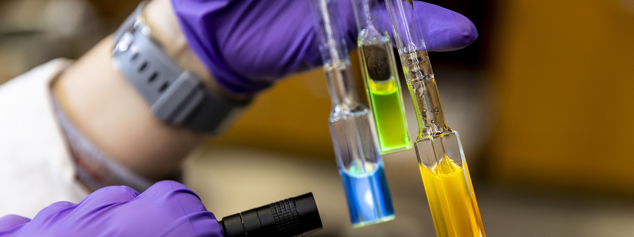 Purple gloved hands hold colorful liquid in scientific glassware while shining a line through the liquid