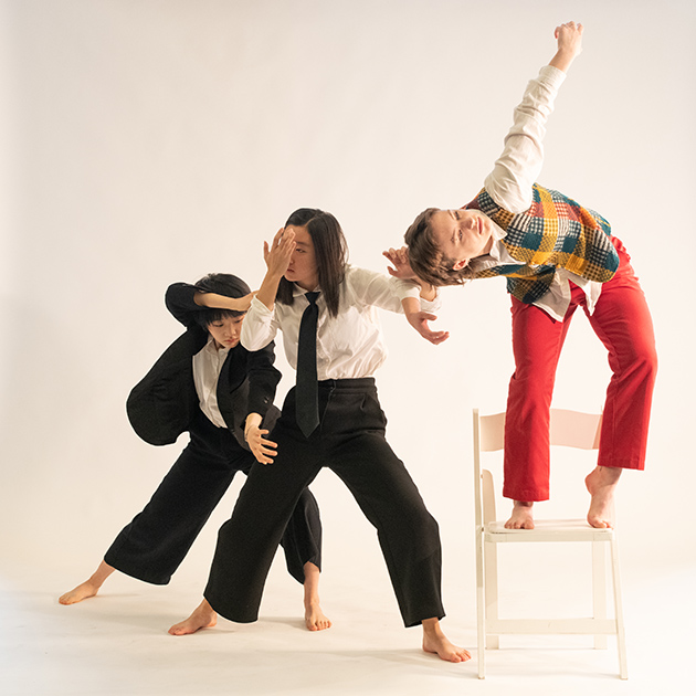 Three dancers performing. One is standing on a chair, two are standing beside her on the floor.