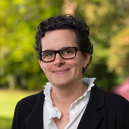 Headshot of Helen Oland wearing glasses and black and white top, set against a blurry tree lined background
