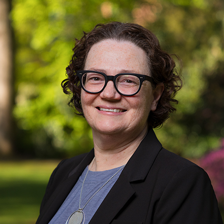 Headshot of Mindy Norris wearing glasses and black jacket, with blurred trees in the background