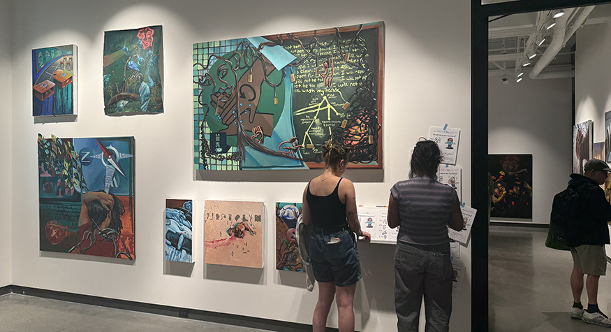 Two students stand beside a gallery wall of artworks by Kyra Wolfenbarger.