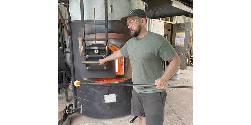 Sean O'Neill holds a pipe with a molten yellow/orange ball of glass on the end.