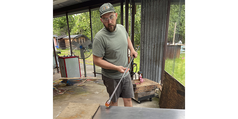 Sean O'Neill holds a glass pipe with hot glass at one end and rolls the glass on a smooth metal surface.
