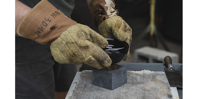 Hands wearing heatproof gloves press a heated glass bowl into other glass that's in a metal mold. 