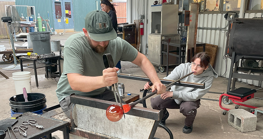 in the studio, one glassblower blows into a blowpipe to force air into a ball of hot glass at the other end of the pipe. The other glassblower shapes the hot glass into sphere. 