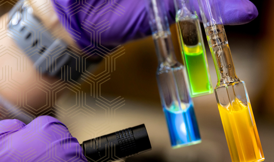 Two hands wearing purple gloves hold pipettes with colorful liquid as the research shines a light through the liquid inside.