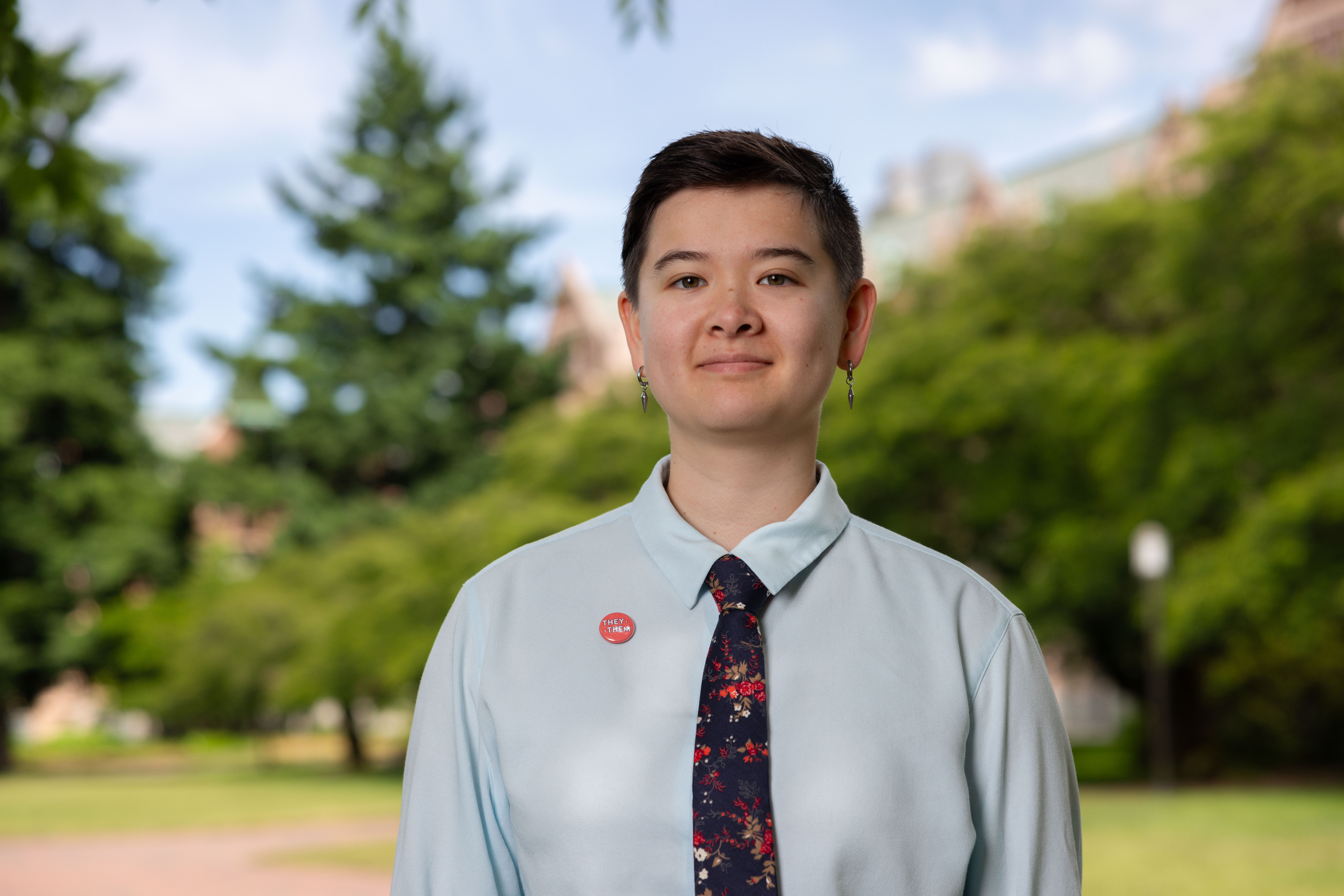 A person with short hair and a tie stands before lunch green trees and lawn.