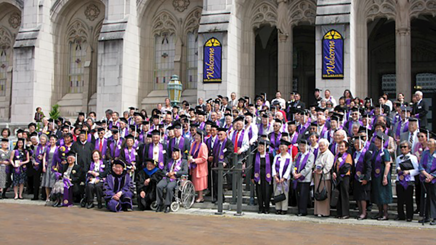 A group of people in front of the Suzzallo library, smiling wearing Husky purple, clutching diplomas