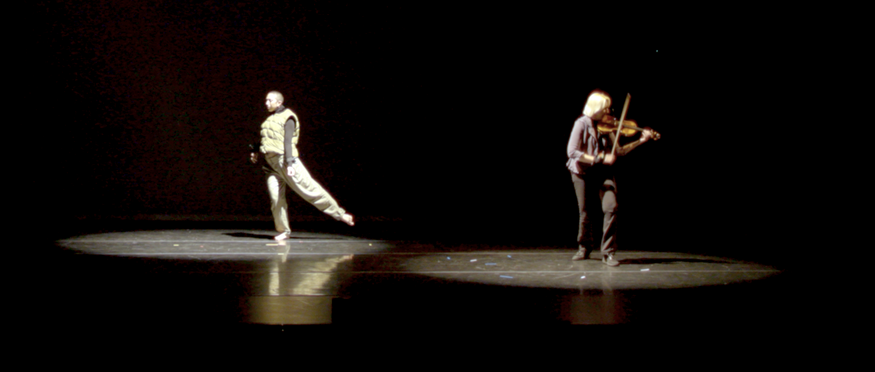 A dancer and violinist perform on a mostly dark stage.