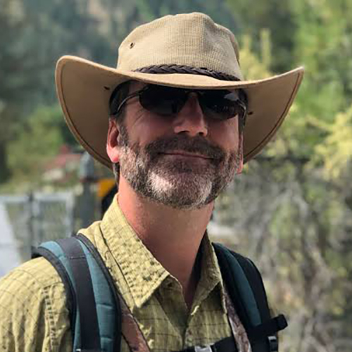 Derek Sheffield in the outdoors, wearing a backpack, with trees in the background.