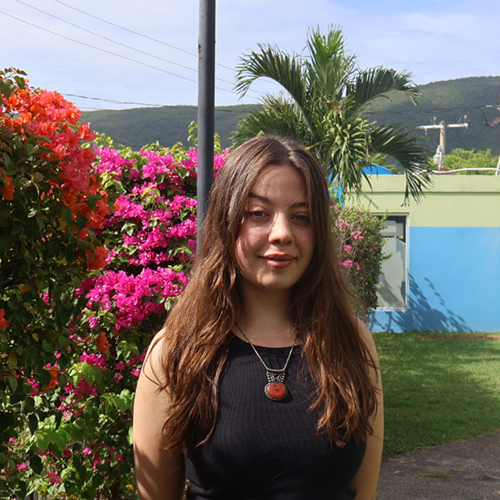 Julia Hayes-Siltzer in Jamaica, with a palm tree, flowers, and a bright blue building in the background.