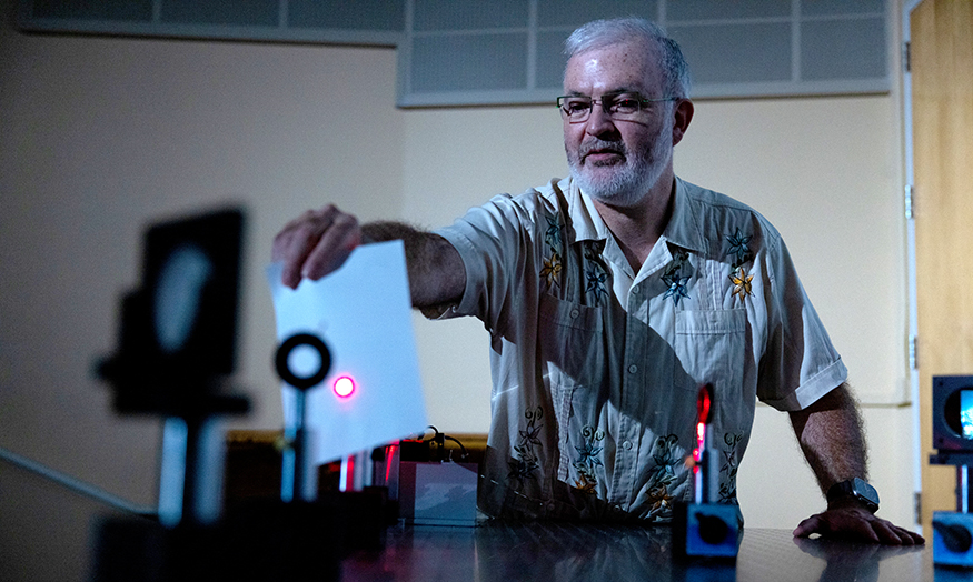 Miguel Morales in a dark room, presenting a demonstration using a pink/red laser. 