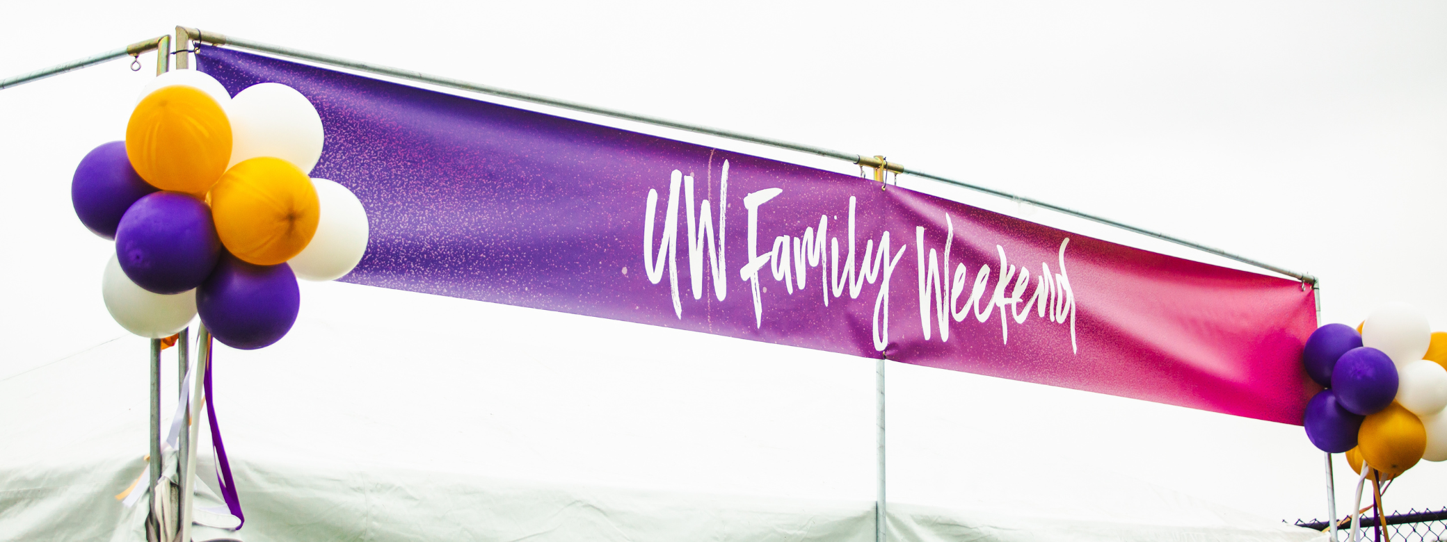 Vibrant purple and pink banner with text that reads: "UW Family Weekend"; A bundle of purple, gold, and white balloons hang on both sides of the banner.