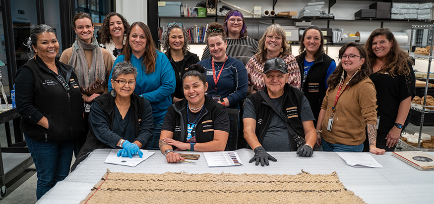 A large group of Coast Salish Wool Weaving Center weavers, with a large woven piece on the table in front of them.