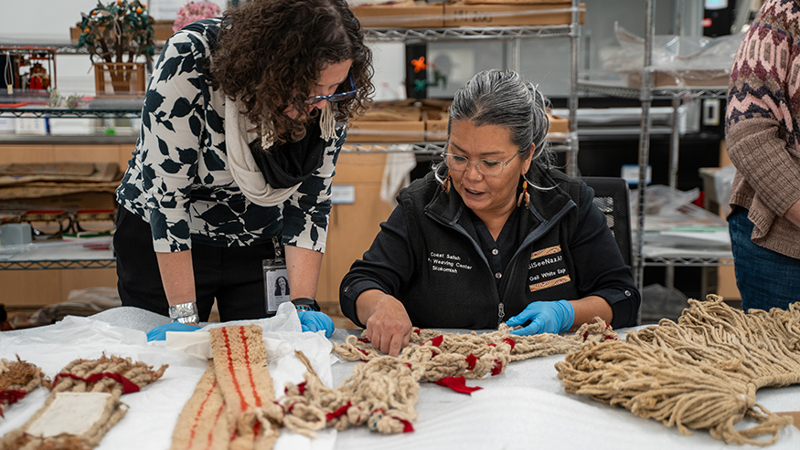 Katie Bunn-Marcuse and Gail White Eagle look at and discuss woven objects spread out on a white table. 